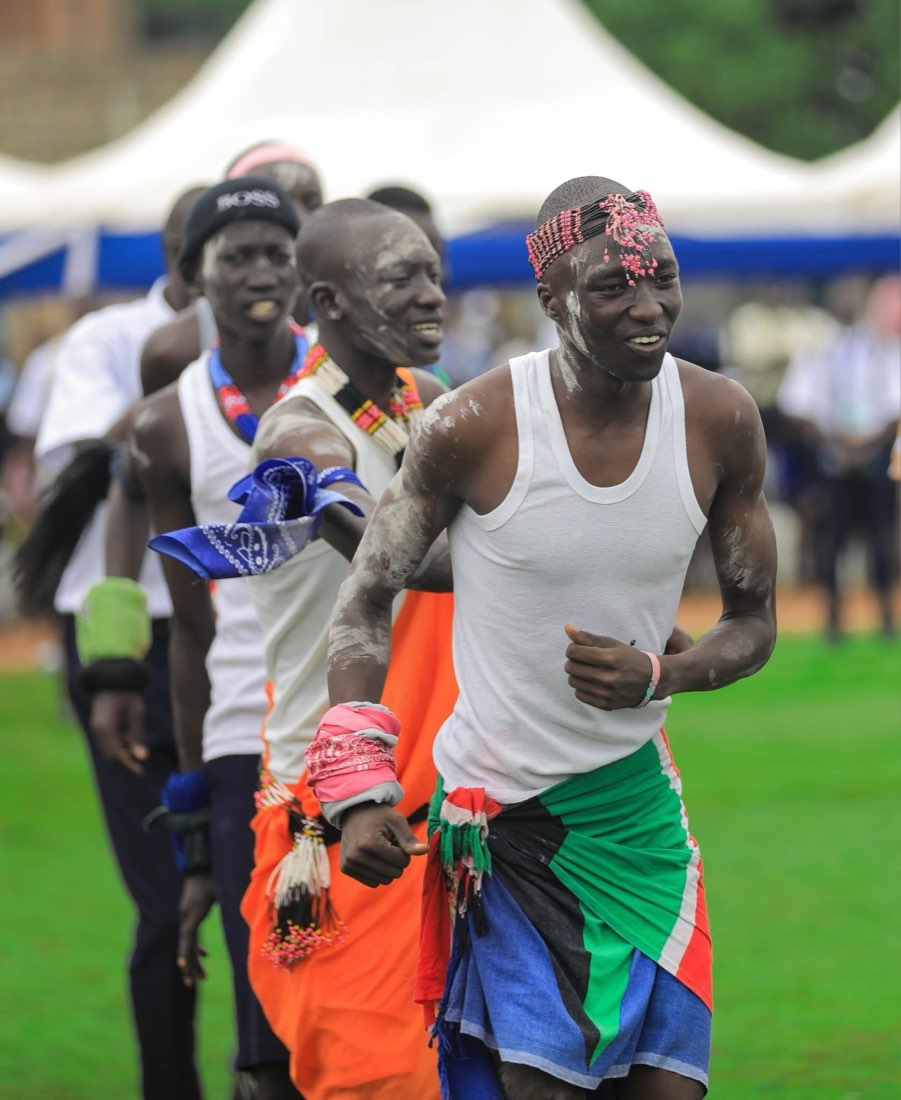Young men in traditional dress dance in a joyful line, one wearing the South Sudanese flag