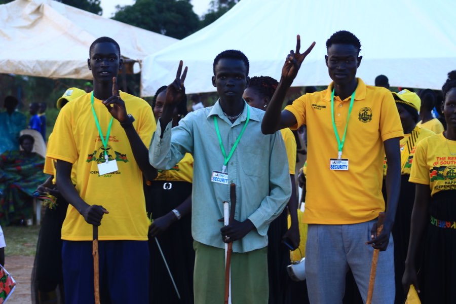 Three young men in event shirts raise peace signs in front of the gathering tents